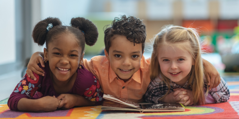 Three children smiling and lying on a colorful carpet with an open book in front of them.