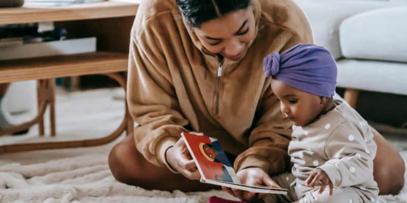 Woman reading a book to a child on a rug in a living room.