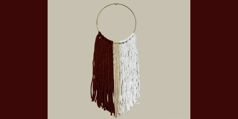 A circular hoop with hanging strands of dark red, tan, and off-white yarn against a beige backdrop.