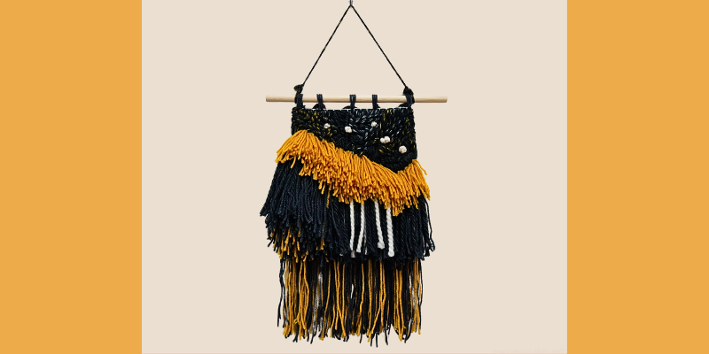 Woven wall hanging with black, mustard yellow, and white yarn, suspended from a wooden dowel.
