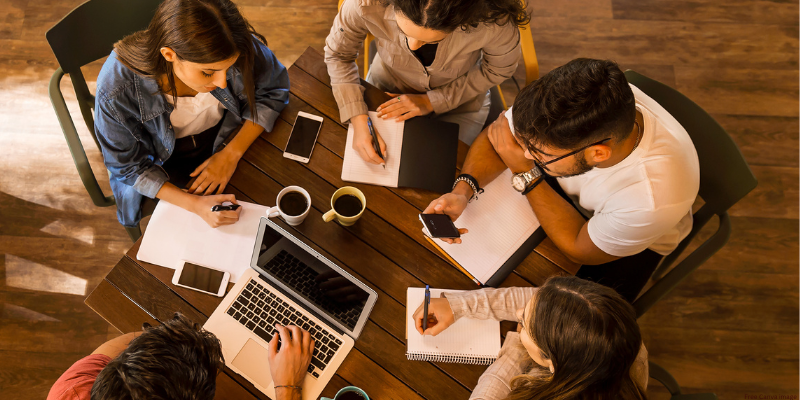 Group of five people in a meeting around a table with a laptop and notebooks.