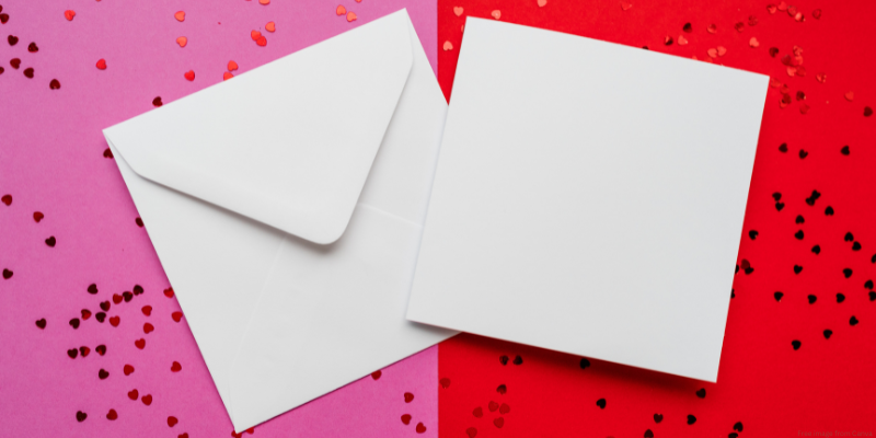 A white envelope and blank square card on a pink and red background with scattered heart-shaped confetti.