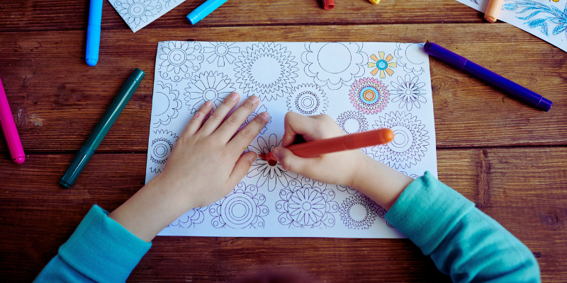 Child coloring floral patterns with markers on a wooden table.