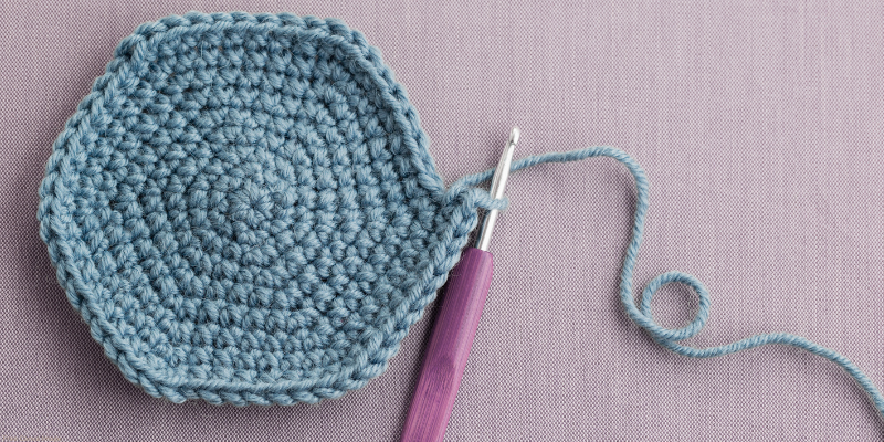 A hexagonal crochet piece with a blue yarn and purple-handled hook on a pink fabric background.