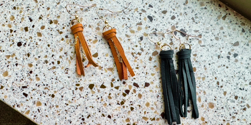 Two pairs of tassel earrings, one brown with gold chain and one black, on a speckled surface.