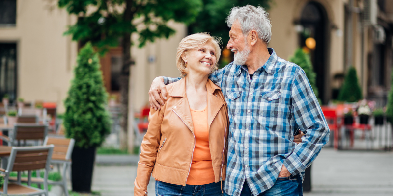 An older couple walking arm in arm, smiling at each other in an outdoor urban setting.