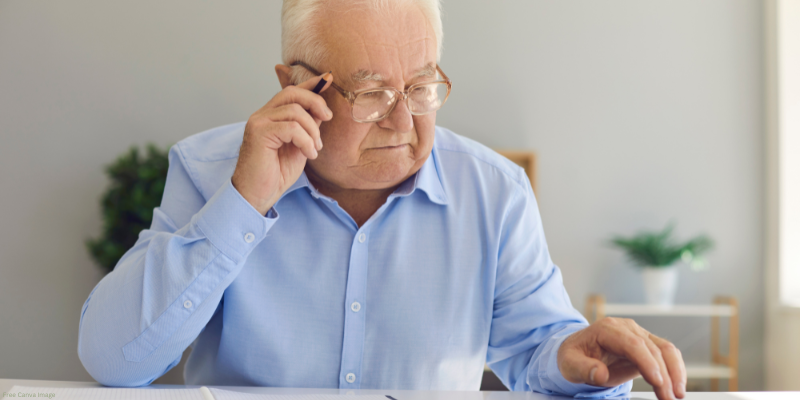 Elderly man in a blue shirt holding a pencil and looking at documents on a table.