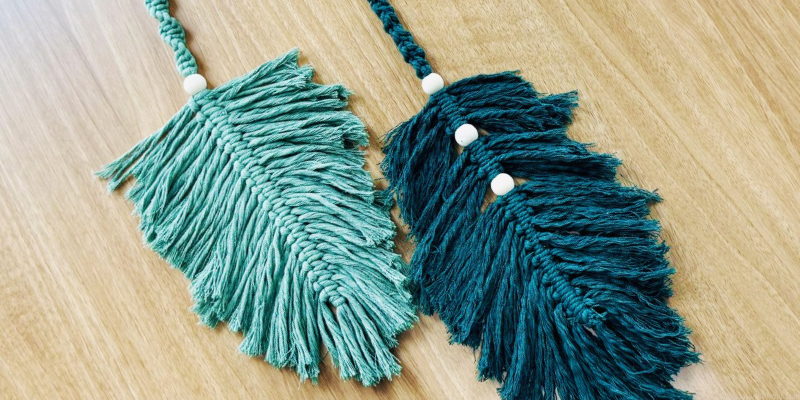 Two macramé leaves on a wooden table, one light green with a bead, the other dark teal with three beads.