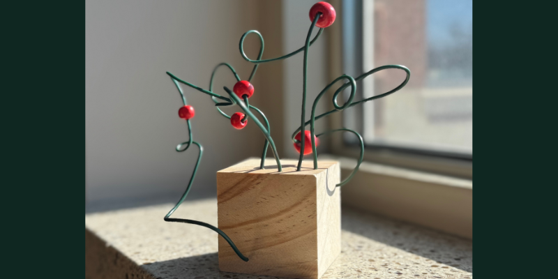 Abstract wire sculpture with red spheres on a wooden base by a sunny window.