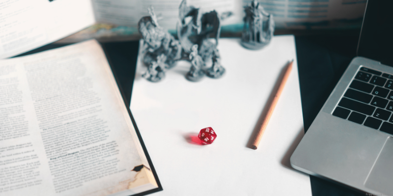 close up of mini figurines, dice, a laptop, paper and pencil, and an open book