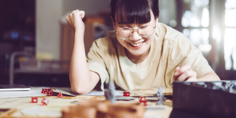 Person smiling while playing a dungeons and dragons game with red dice and figurines.