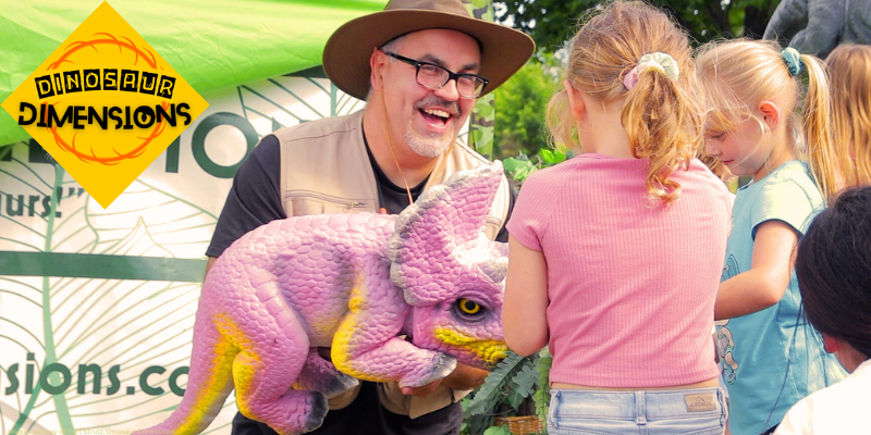 A man holding a pink and yellow toy dinosaur with logo in corner that reads "Dinosaur Dimensions"
