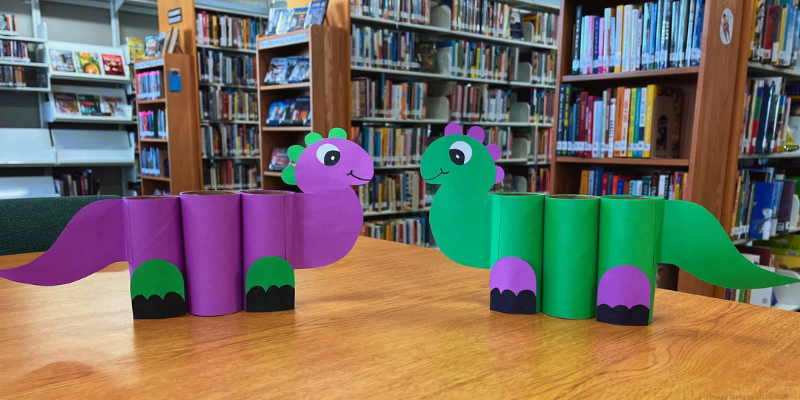background of library bookshelves with a table showing two paper dinosaur pencil holders facing each other