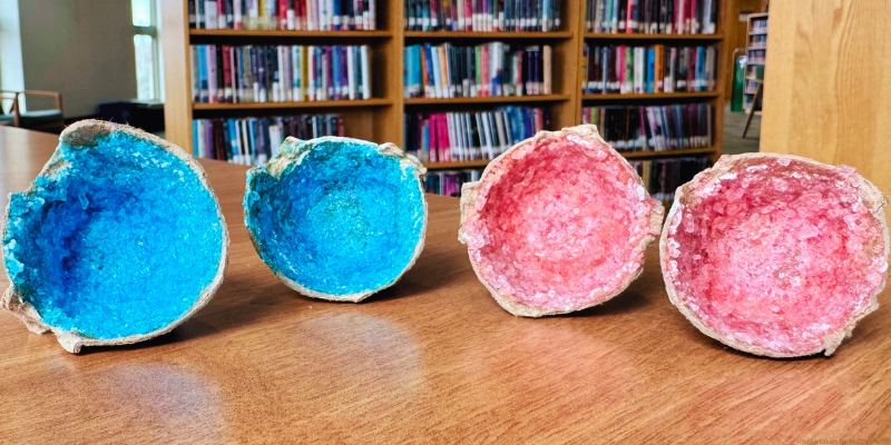 Four egg carton cups showing geode-like vibrant blue and pink interiors on a wooden table, with bookshelves in the background.
