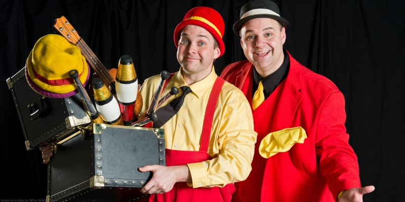 two men in red and yellow baggy outfits smiling and holding a box of props