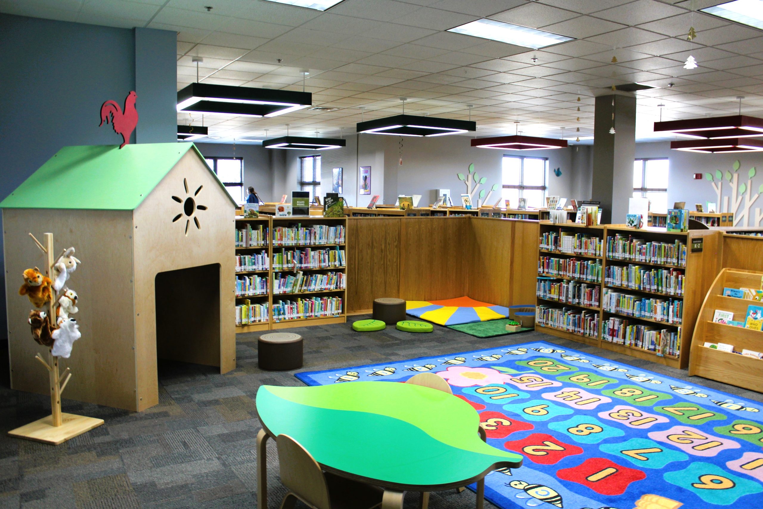 Children's library area with a playhouse, plush toys, colorful number carpet, and book-filled shelves.