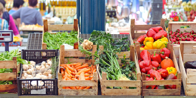 A market stall with crates of garlic, carrots, leafy greens, bell peppers, and chili peppers.
